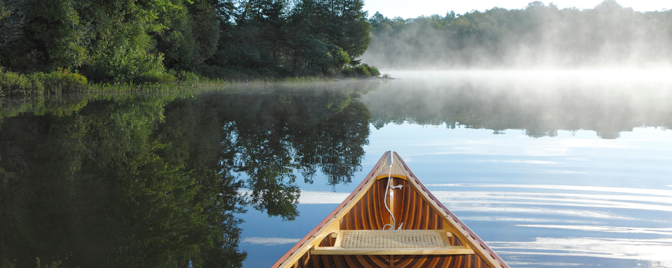 Beyond the bow of a canoe can be seen a misty lake edged with lush green trees.